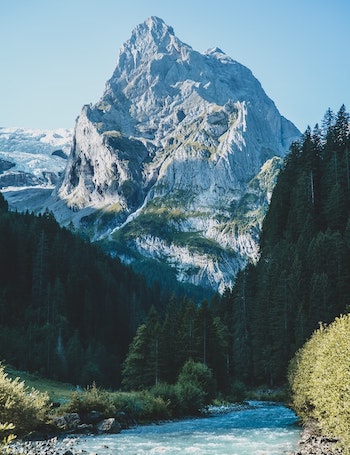 A towering, jagged mountain peak rises above a forest and a river.